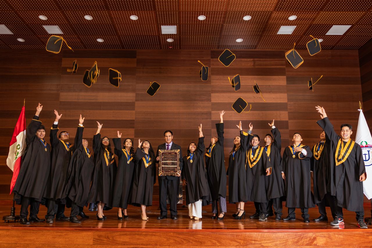 Graduates celebrating by tossing caps indoors in Tacna, Perú, marking their achievement with joy.