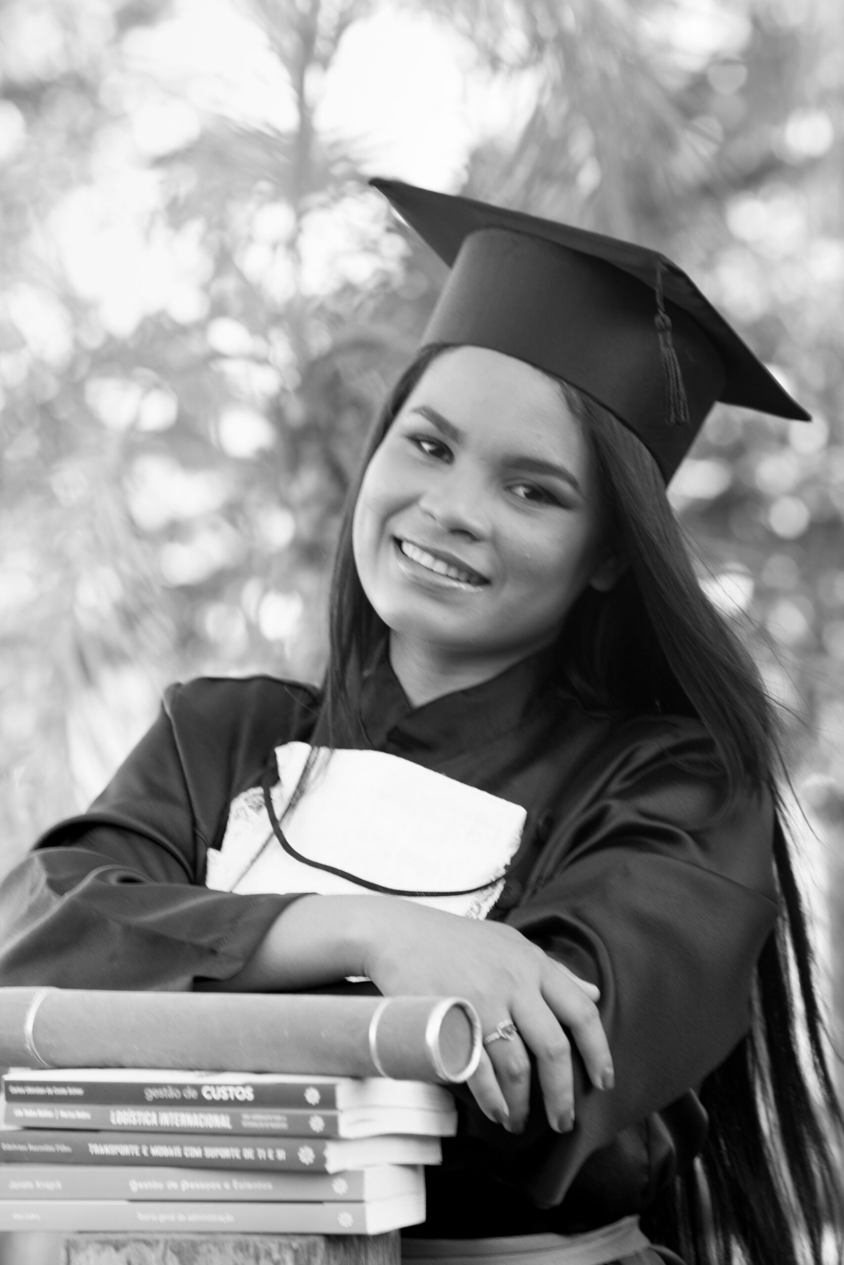 Smiling graduate in cap and gown posing with books in an outdoor setting.